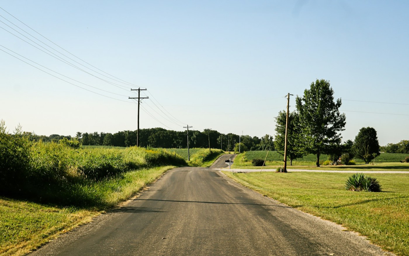 A paved rural road with grass shoulders and wooden utility poles running along both sides.